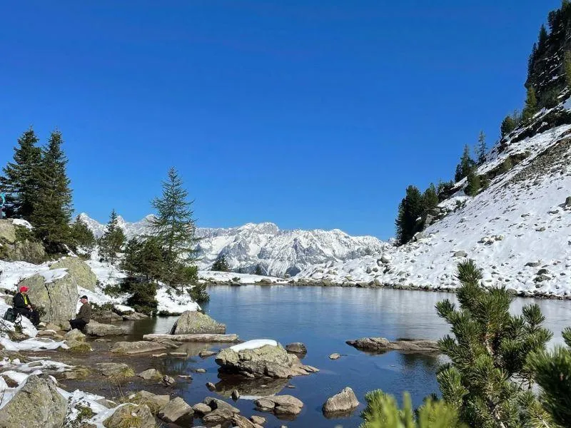 Mountain lake surrounded by snowy rocks and trees, with two people sitting on distant rocks enjoying the view
