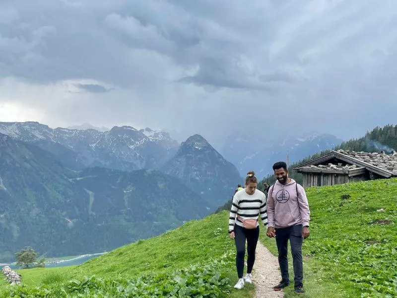 Couple holding hands on a mountain hiking trail with panoramic views of a lake and mountains behind them