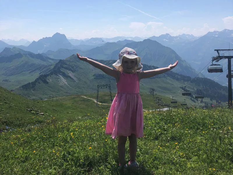 Little girl standing in a green alpine meadow with chairlift and mountains in the background during summer