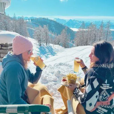 Two women enjoying Radler beer and fries in snowy Alpine sun, relaxing in lounge chairs with blankets after skiing
