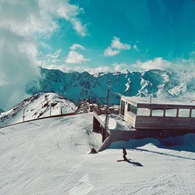 Scenic high-Alpine view with modern wooden ski hut, single skier on empty snowy slope, and vivid blue sky in the European Alps