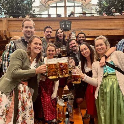 Group of friends standing on benches in an Oktoberfest beer tent, holding up large Bavarian beer steins and cheering happily, celebrating the festive Munich Oktoberfest spirit.