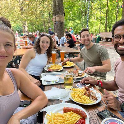 Friends sitting in a Munich Biergarten on a sunny day, drinking large Bavarian beers and eating traditional food, surrounded by green trees and lively atmosphere.