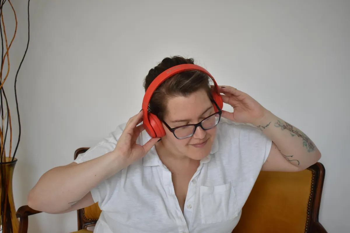 Cheryl sits on a yellow fabric antique settee. They are wearing red headphones and a white tshirt