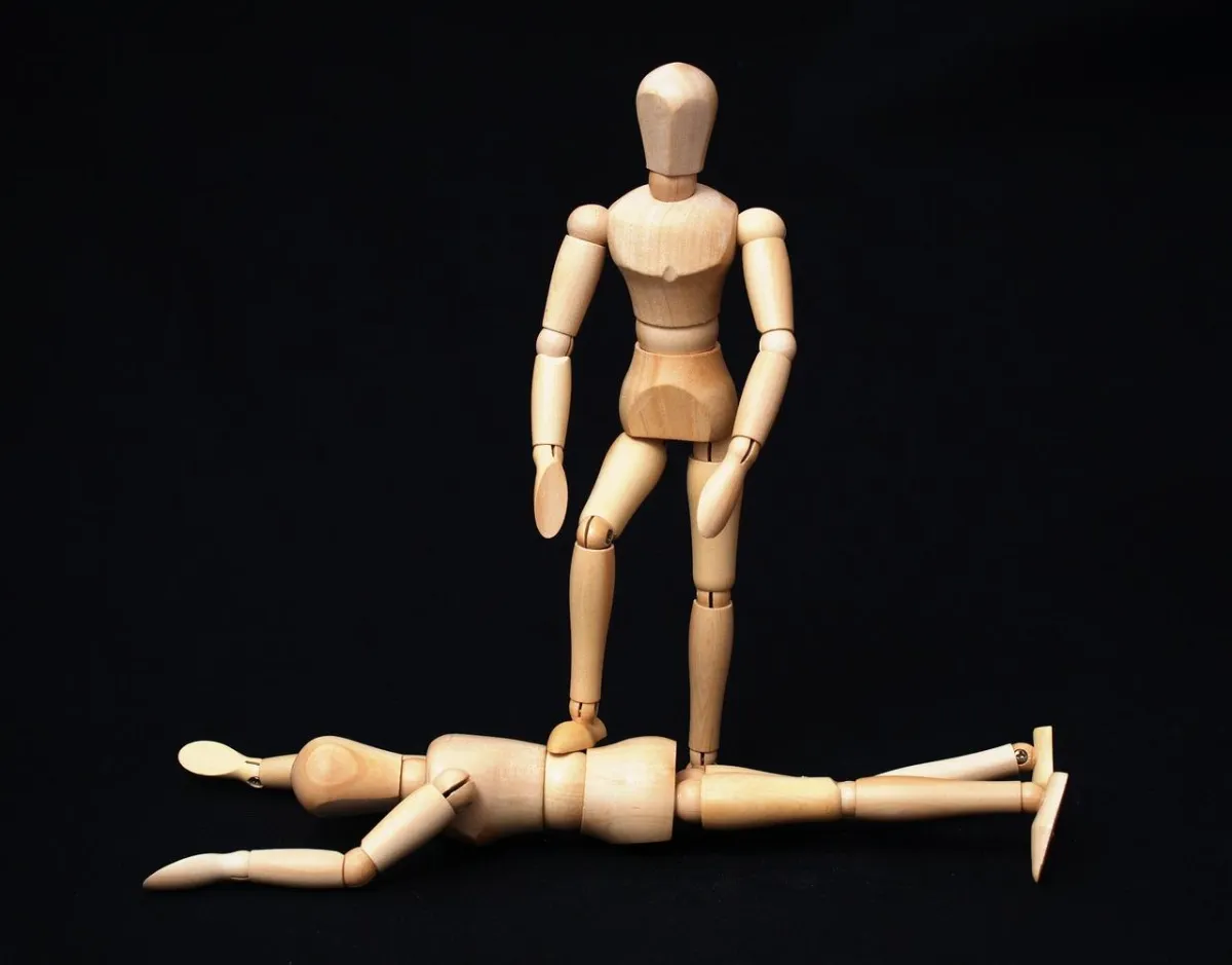 a wooden stick figure of a man standing with one foot on a woman's back as she is face down on the floor