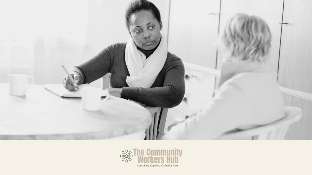 A woman sits at a table taking notes while listening attentively to another person seated opposite her. The black-and-white image shows a calm, supportive conversation in a domestic or office setting, with cups on the table. The Community Workers Hub logo appears at the bottom of the image.