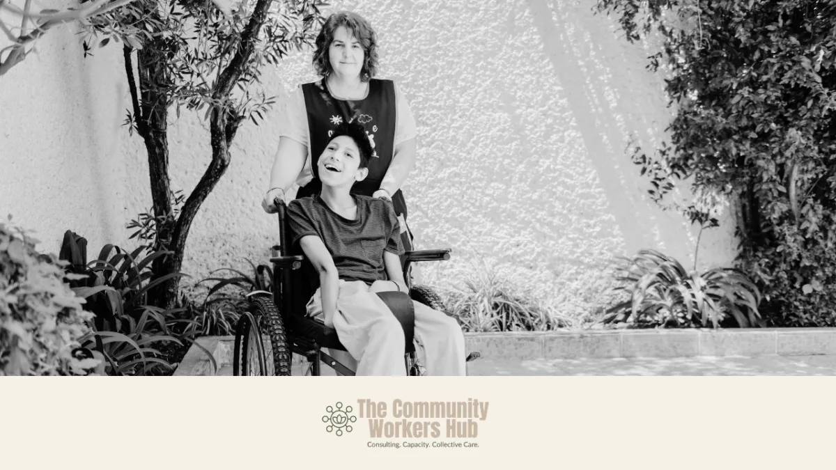 A black and white image of a young person in a wheelchair. A adult carer is stading behind the wheelchair. They are outside with a wall and plants in the background.