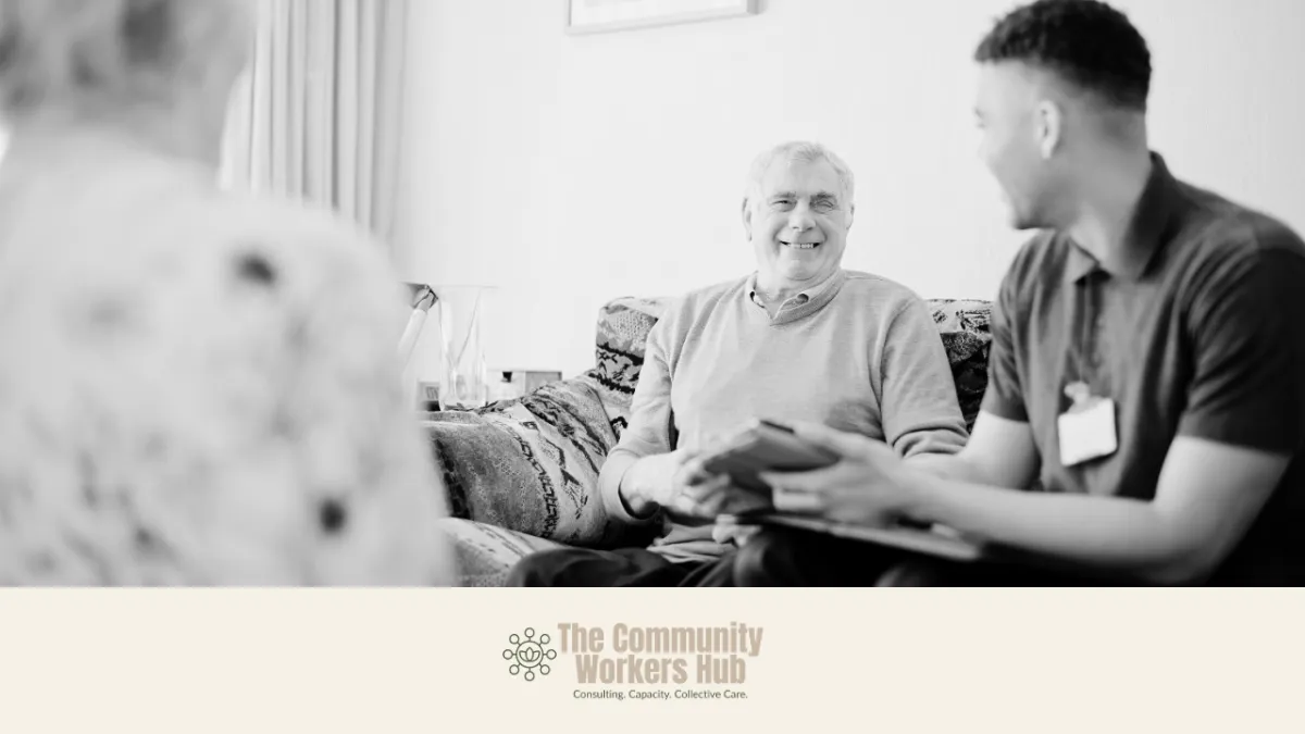An older man sits on a couch smiling and talking with a support worker who is holding a tablet or clipboard, while another person is blurred in the foreground. The scene suggests a friendly, supportive conversation in a home setting. The Community Workers Hub logo appears at the bottom of the image.