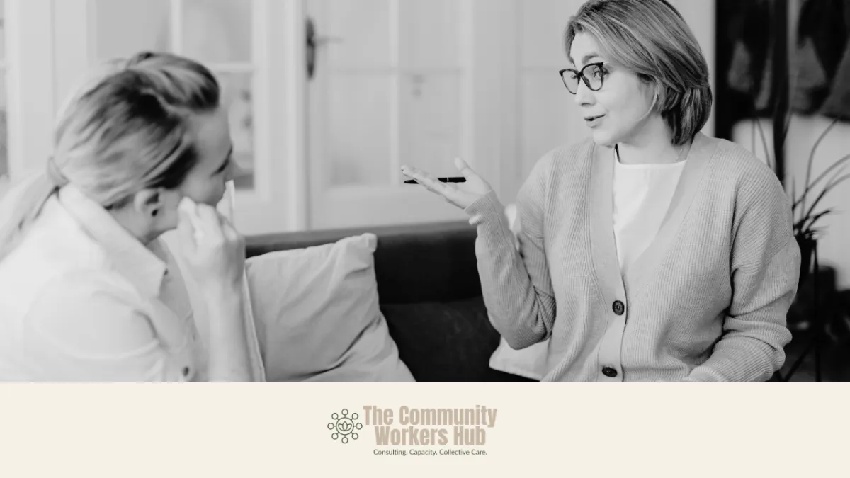 Two women sit on a couch engaged in conversation, with one woman gesturing as she speaks and the other listening attentively. The black-and-white image shows a supportive, reflective discussion in a comfortable indoor setting. The Community Workers Hub logo appears at the bottom of the image.