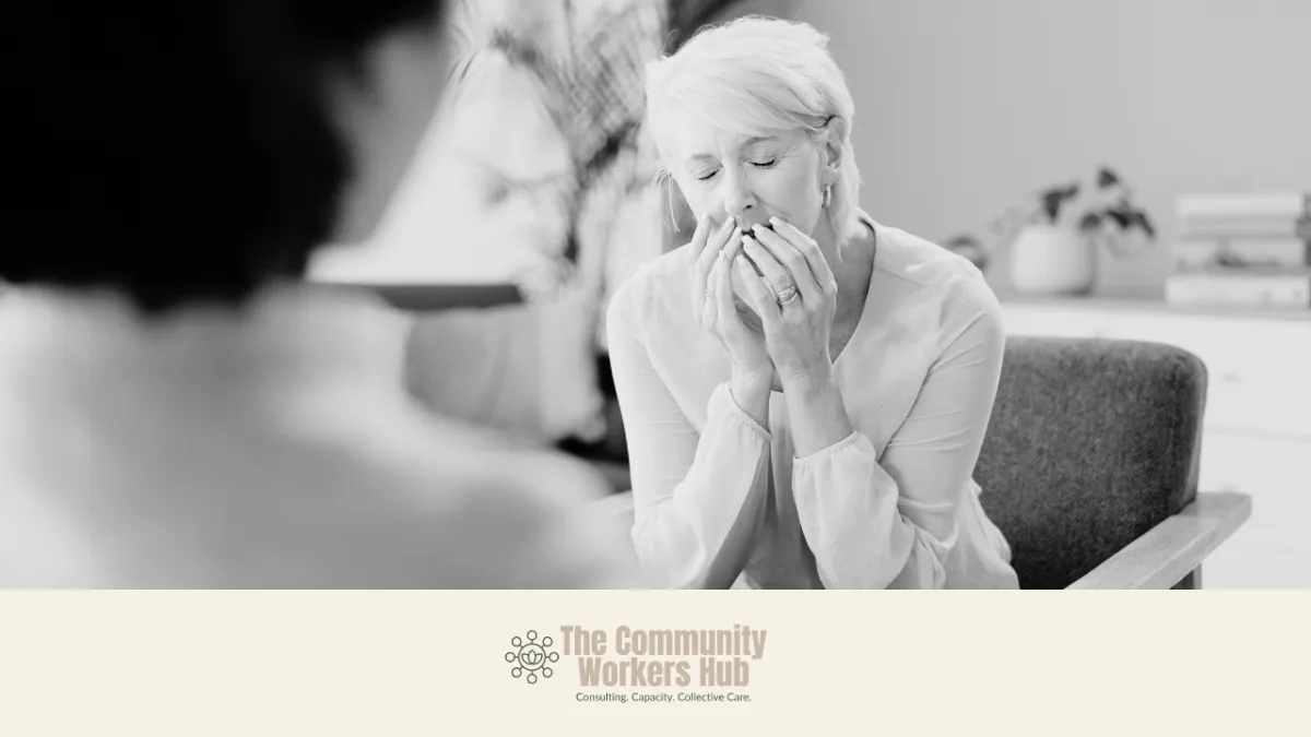 An older woman sits indoors with her hands covering her mouth, eyes closed, appearing deeply distressed or emotional. Another person is blurred in the foreground, suggesting a supportive conversation. The black-and-white image conveys vulnerability, grief, or complex trauma. The Community Workers Hub logo appears at the bottom of the image.