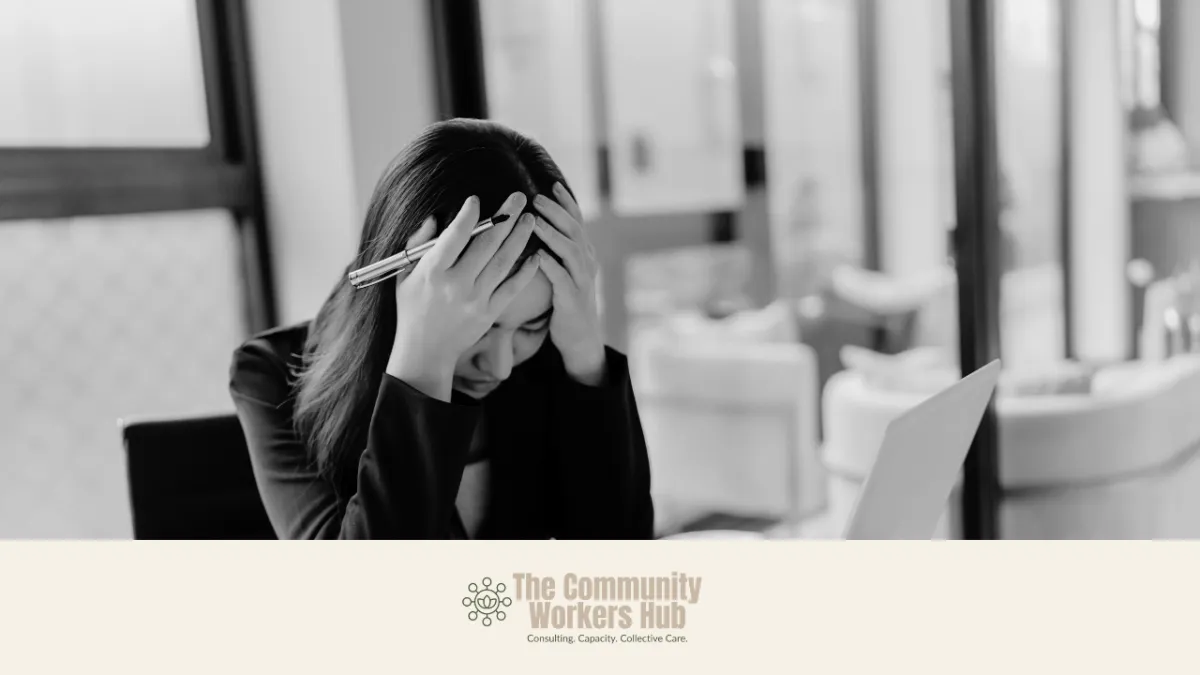 A woman sits at a desk with her head in her hands, holding a pen, looking distressed while facing an open laptop. The black-and-white image conveys stress, overwhelm, or system pressure in a work setting. The Community Workers Hub logo appears at the bottom of the image.