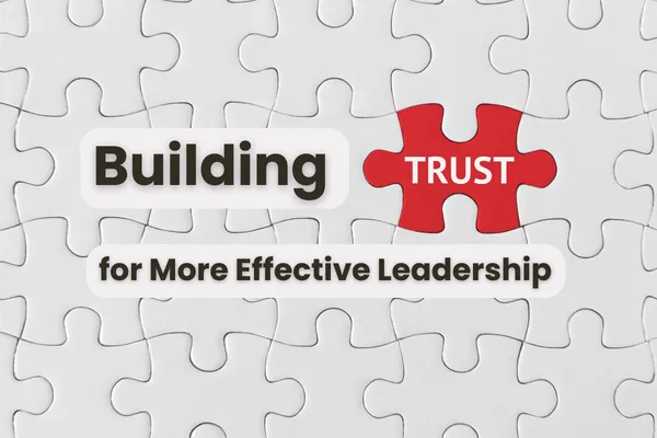 a field of white puzzle pieces in which one is red with white text overlaid, reading, "TRUST," and around which are floating words that spell out the blog article title: "Building Trust for More Effective Leadership," visually incorporating the puzzle piece text into the title