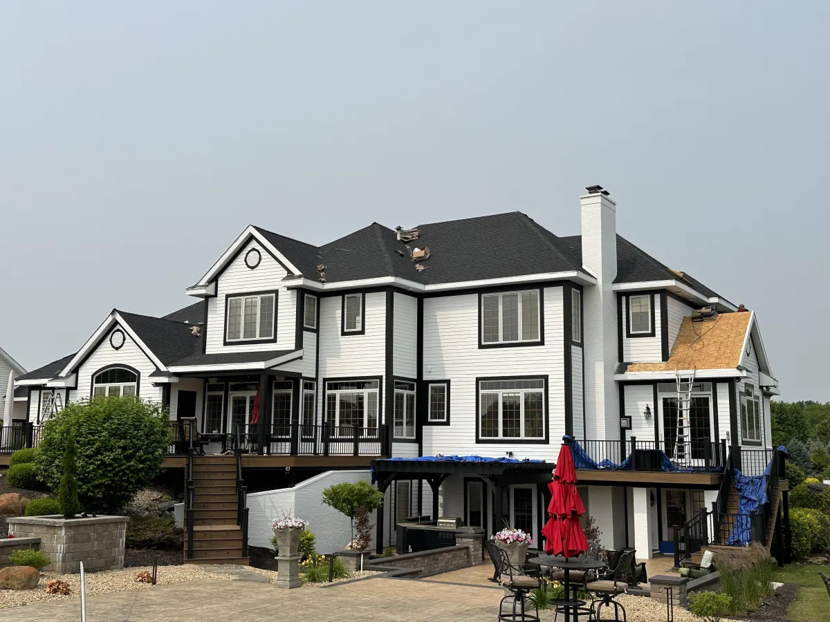 Large two-story white and black house undergoing partial roof replacement. The home features multiple gables, black-trimmed windows, a large deck with red umbrellas, and a visible section of exposed roof decking with underlayment and a ladder. Blue tarps cover parts of the roof and deck during the construction process.