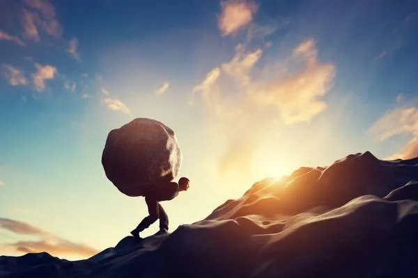a man climbing a mountain while carrying a boulder in his back 
