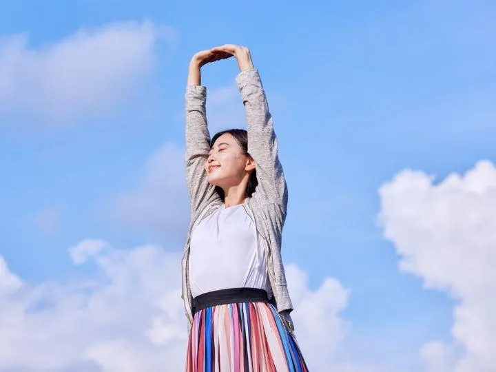 Woman standing outdoors under a blue sky, stretching with arms raised overhead, eyes closed, looking peaceful and relaxed