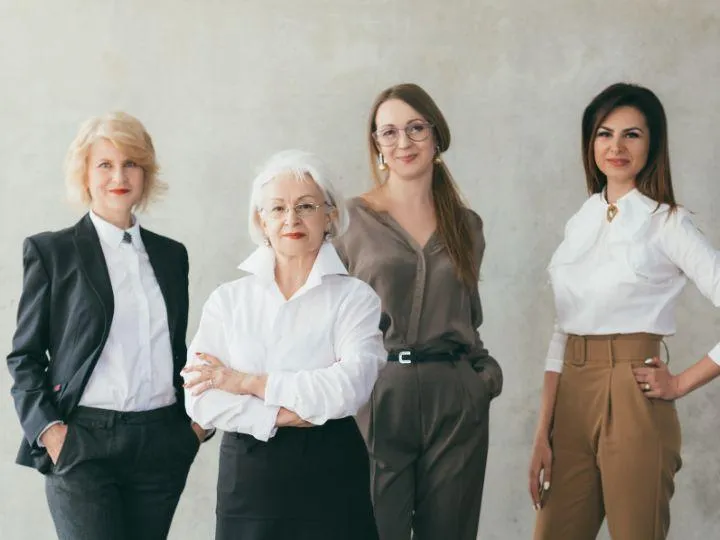 Four professional women of different ages stand together in business attire, smiling confidently against a light, textured wall.