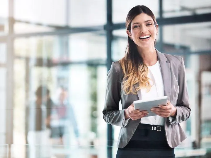 Confident businesswoman in a gray blazer holding a digital tablet and smiling in a modern office setting