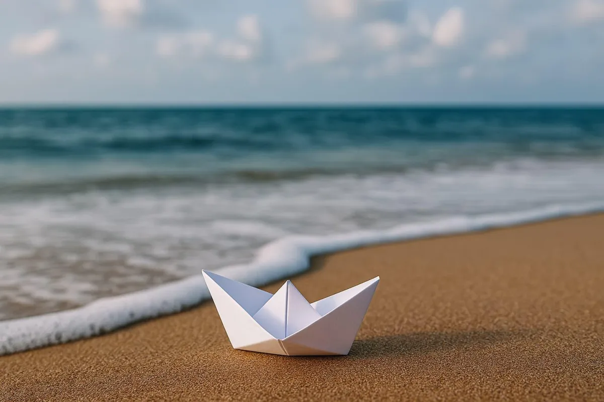 A small white paper boat rests on the golden sand at the edge of the ocean