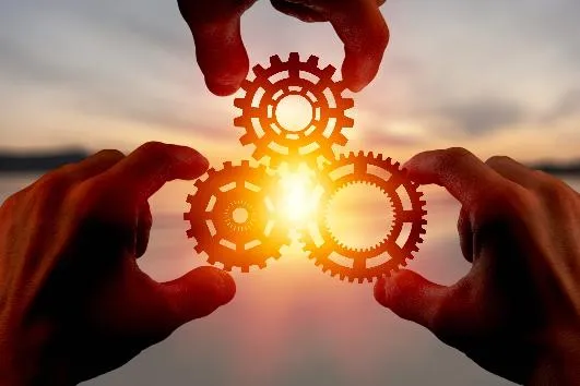 Three hands holding gears in a triangle shape with a sunrise behind it. Business. By BillionPhotos.com