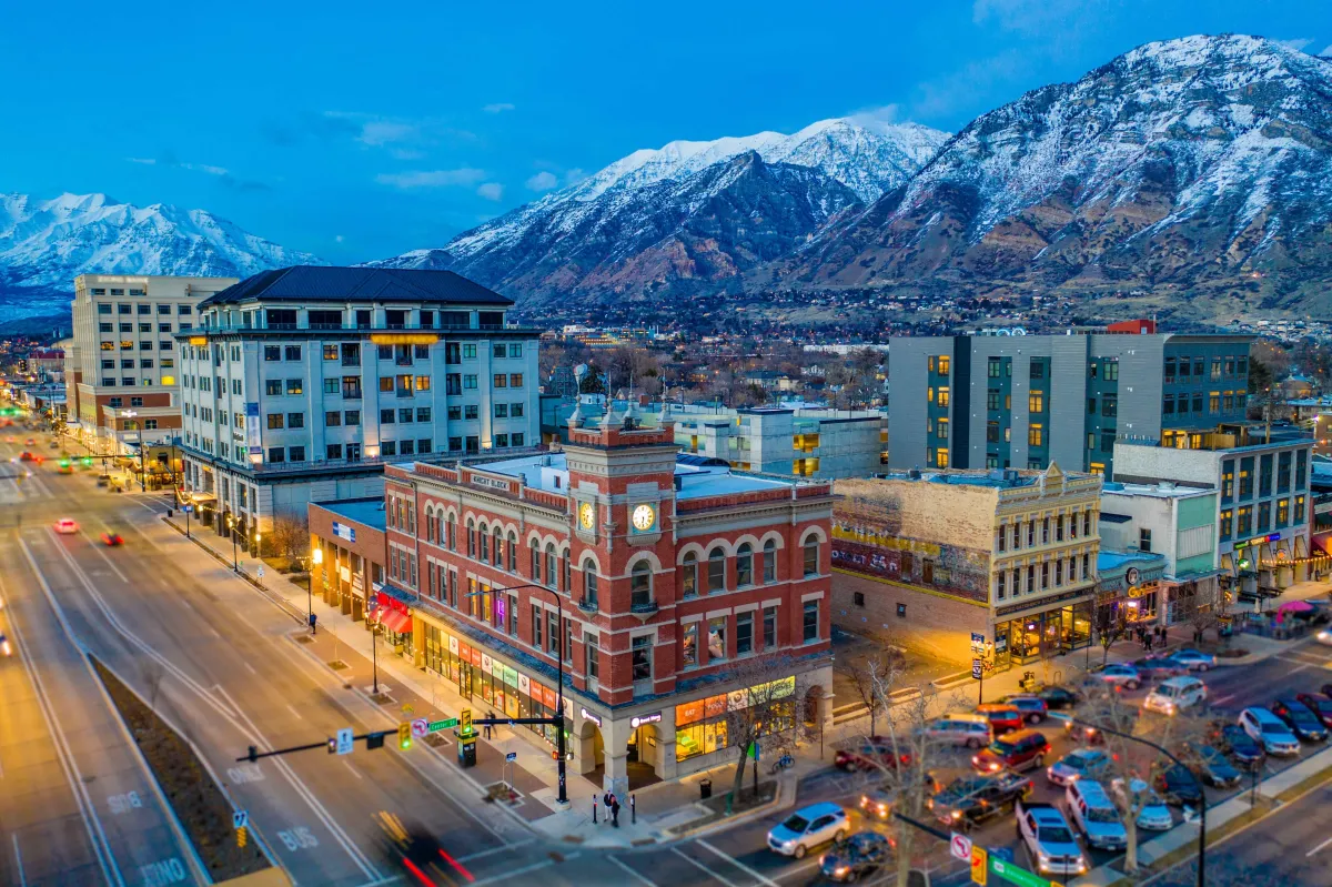 A picturesque winter scene of Downtown Provo, Utah. The image showcases a serene, snow-covered streetscape with buildings lining the sides. The architecture reflects a blend of modern and traditional styles, under a clear, blue winter sky. The snow adds a peaceful, tranquil quality to the urban environment, highlighting the beauty of Provo in the winter season.
