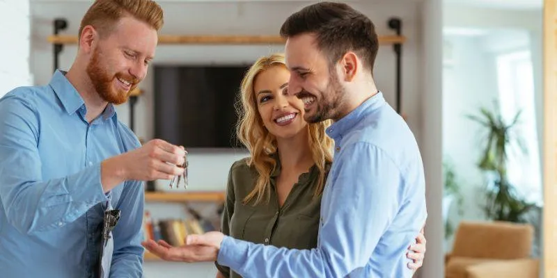 A real estage agent hands house keys to a smiling couple inside a modern, well-lit living room.