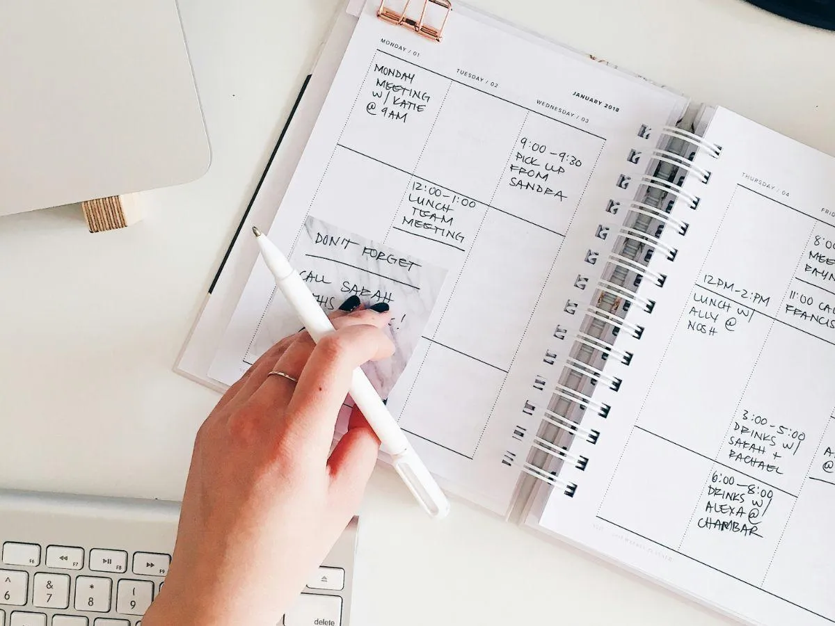A hand holding a pen, writing in an open planner with a week's schedule, including meetings and reminders. A sticky note with "Don't forget, call Sarah" is visible on the page. Every Task Virtual Assistants USA.