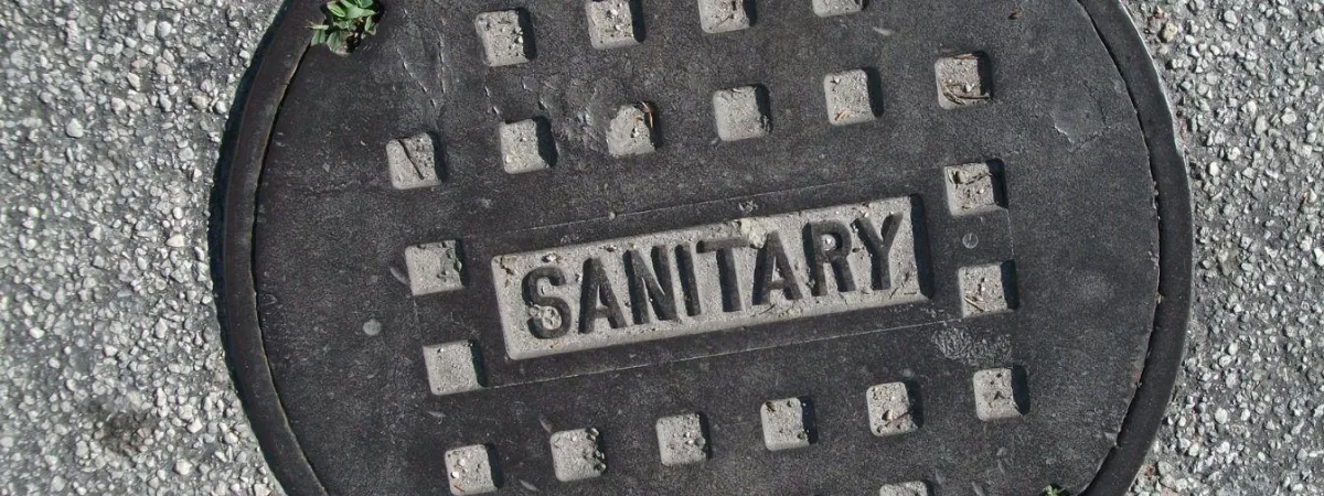 Close-up of a round sanitary sewer manhole cover embedded in asphalt, indicating underground wastewater infrastructure. Essential for municipal sewage systems, proper sewer maintenance helps prevent backups and plumbing issues.
