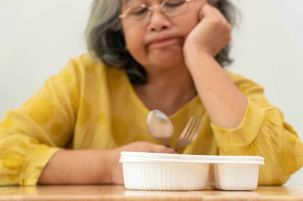 A woman over 50 sits at a table with a plate of food in front of her, looking frustrated and unsure about what to eat