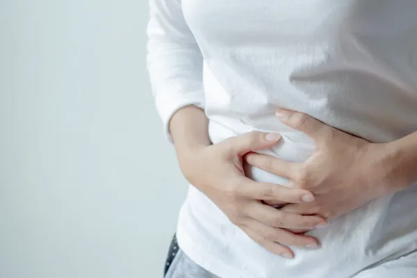 Person clutching their abdomen, indicating stomach pain or digestive discomfort, wearing a white shirt and grey pants.