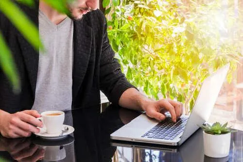 A person working on a laptop at a glass table with a cup of coffee, surrounded by bright green plants