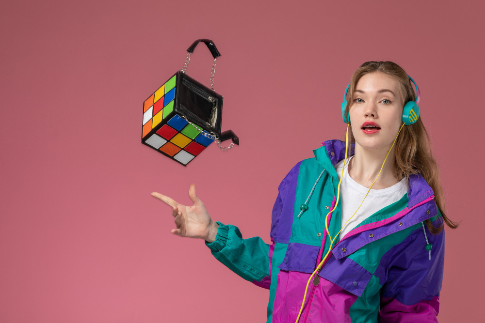 Young woman in retro colorblock windbreaker tossing a Rubik's cube handbag while wearing bright teal headphones, pink background, fun lifestyle and accessories concept