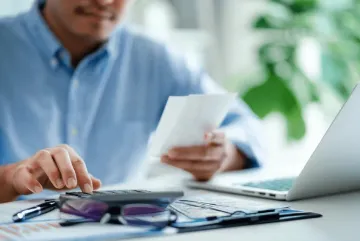 A professional reviews financial documents at a desk, holding a receipt while using a calculator. Surrounded by charts, a laptop, and office tools