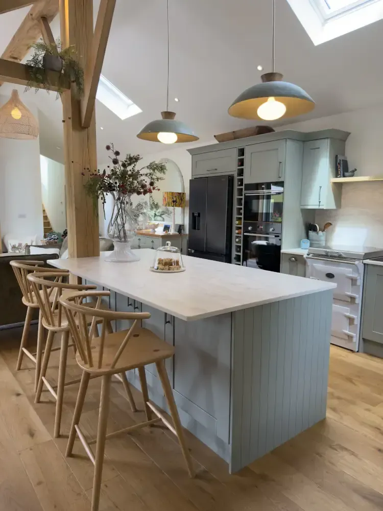 Sage green shaker kitchen with marble island, exposed oak beams, skylights, and natural wood Windsor bar stools in a modern country style design by Blueprint Kitchens