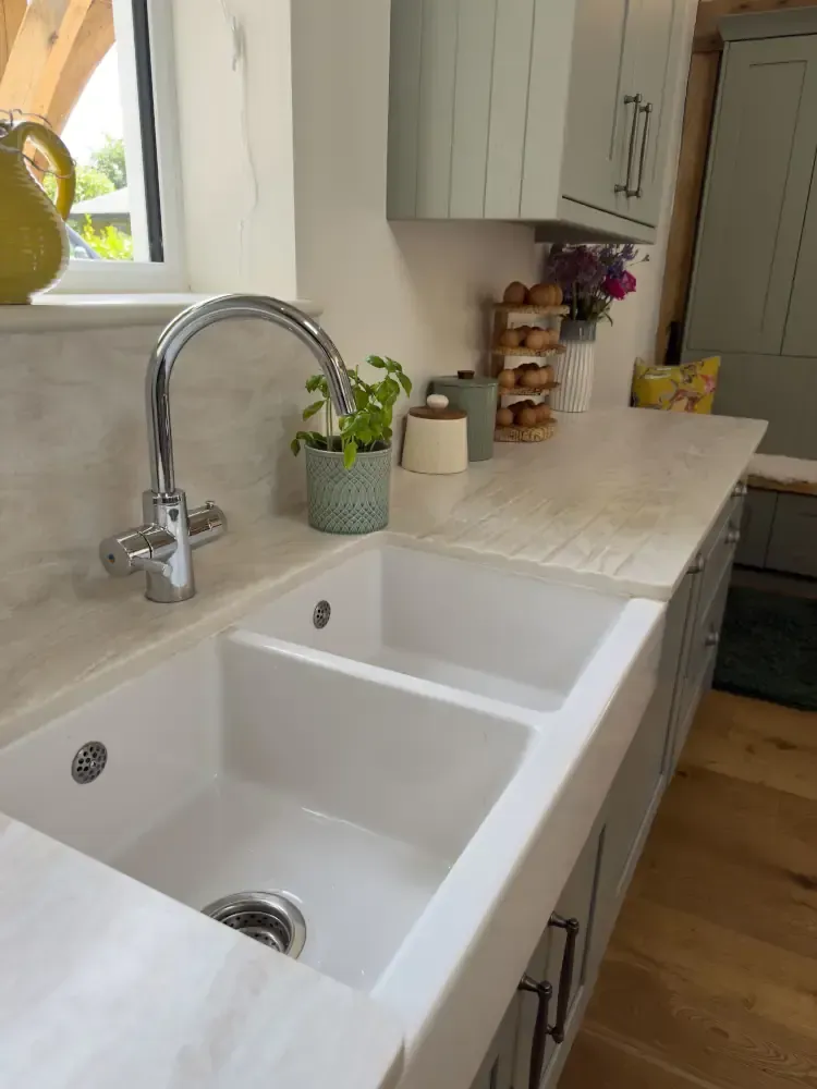White ceramic butler sink with chrome tap, sage green cabinets, marble countertops, and kitchen accessories by Blueprint Kitchens
