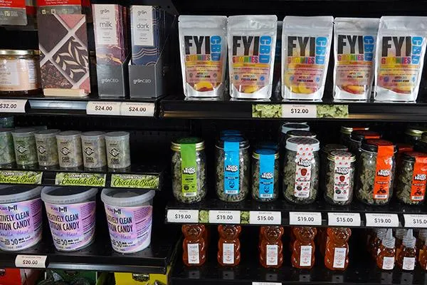 Retail display shelf stocked with various cannabis and CBD products, including gummies, jars of cannabis flower, chocolates, infused honey, and confectionery items with price tags.