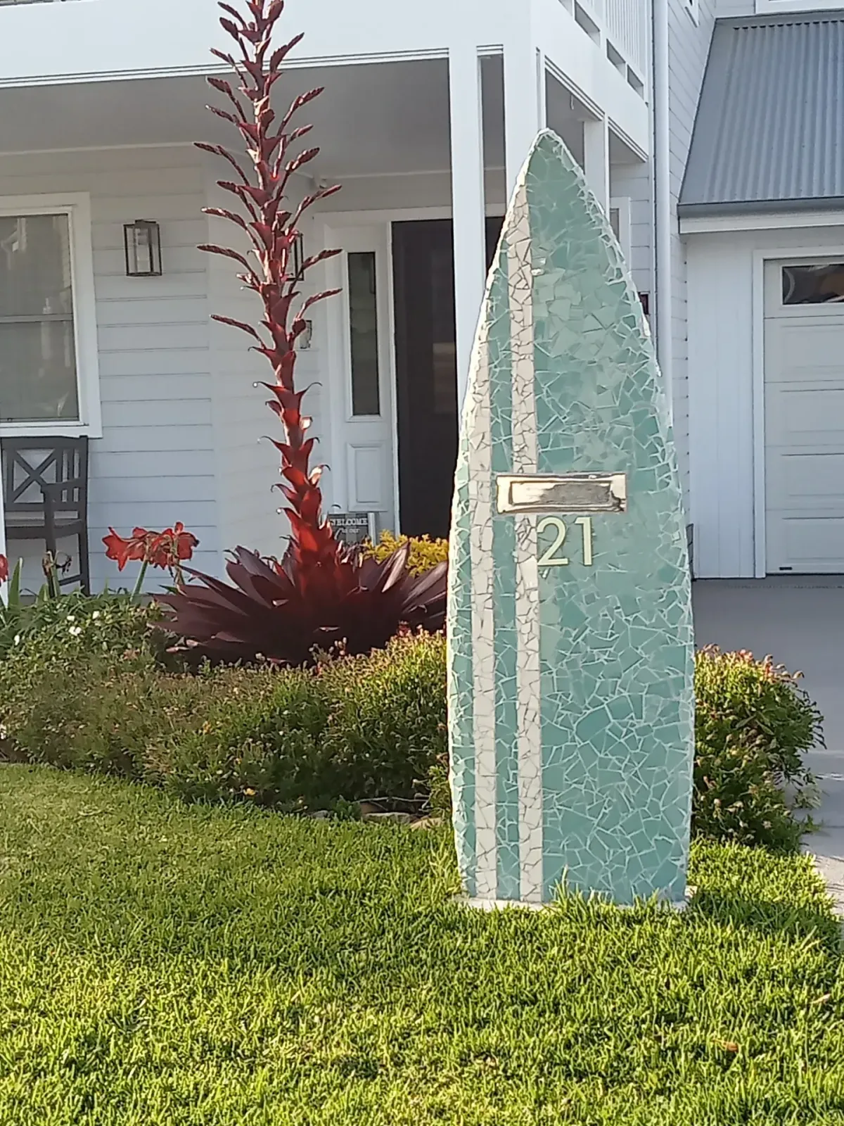 Mint-green mosaic surfboard letterbox with twin white stripes installed in a Hamptons-style front yard