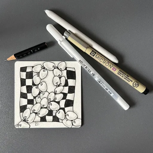 A Zentangle® tile created by Lisa Crow, CZT. The tile is sitting on a table next to a pencil, a Micron 03 pen, a white Gelly Roll pen and a blending stump