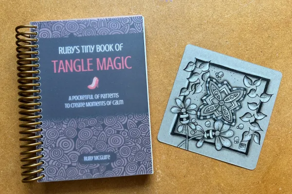 A copy of Ruby's Tiny Book of Tangle Magic sits next to a paper tile with patterns drawn on it from inside the book.