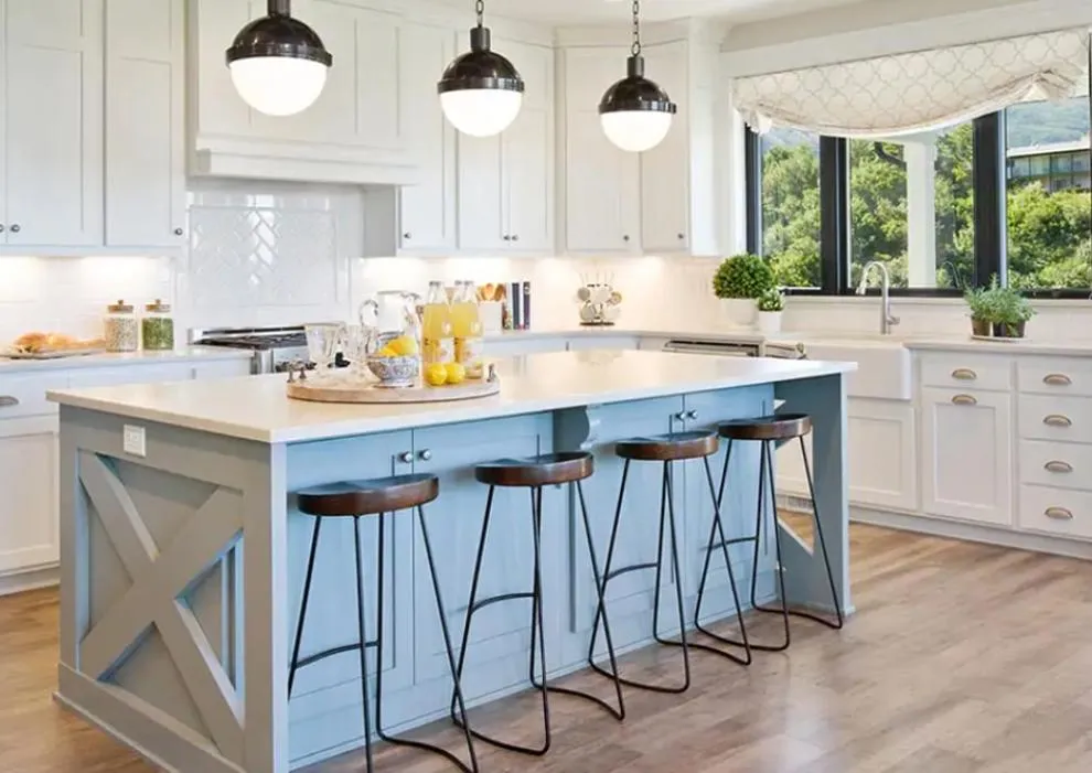 Modern farmhouse kitchen with a light blue island, white cabinets, black pendant lights, and large windows overlooking greenery.