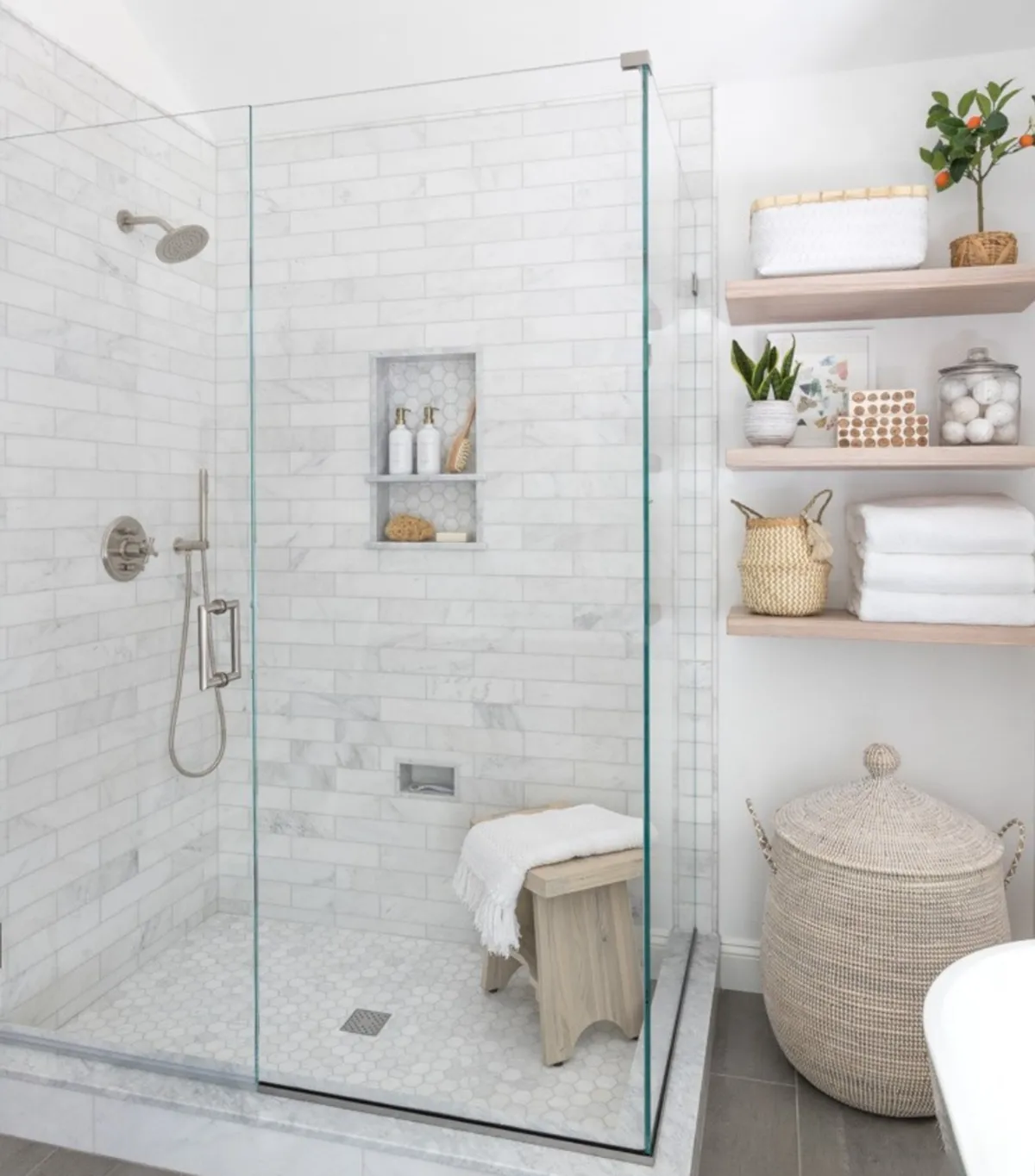 Bathroom with a frameless glass marble-tiled shower, recessed shelf, wooden bench, and open wooden shelving with towels and baskets.