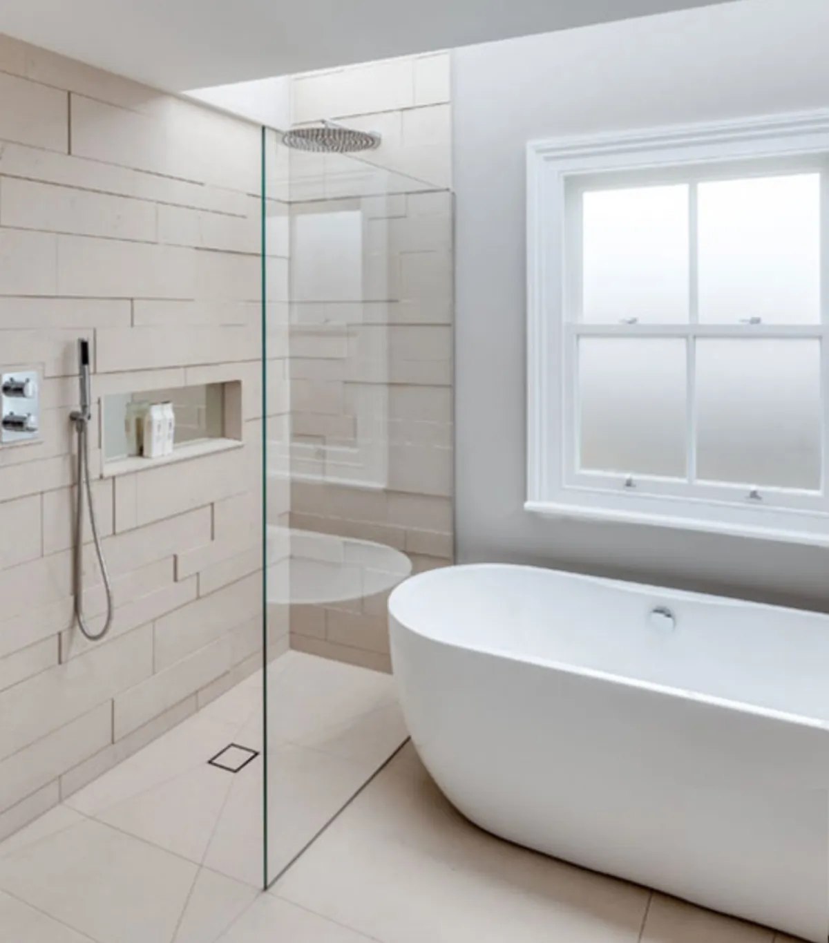 Modern bathroom with a freestanding white bathtub, frameless glass shower, beige tiles, and a rain showerhead next to a frosted window.