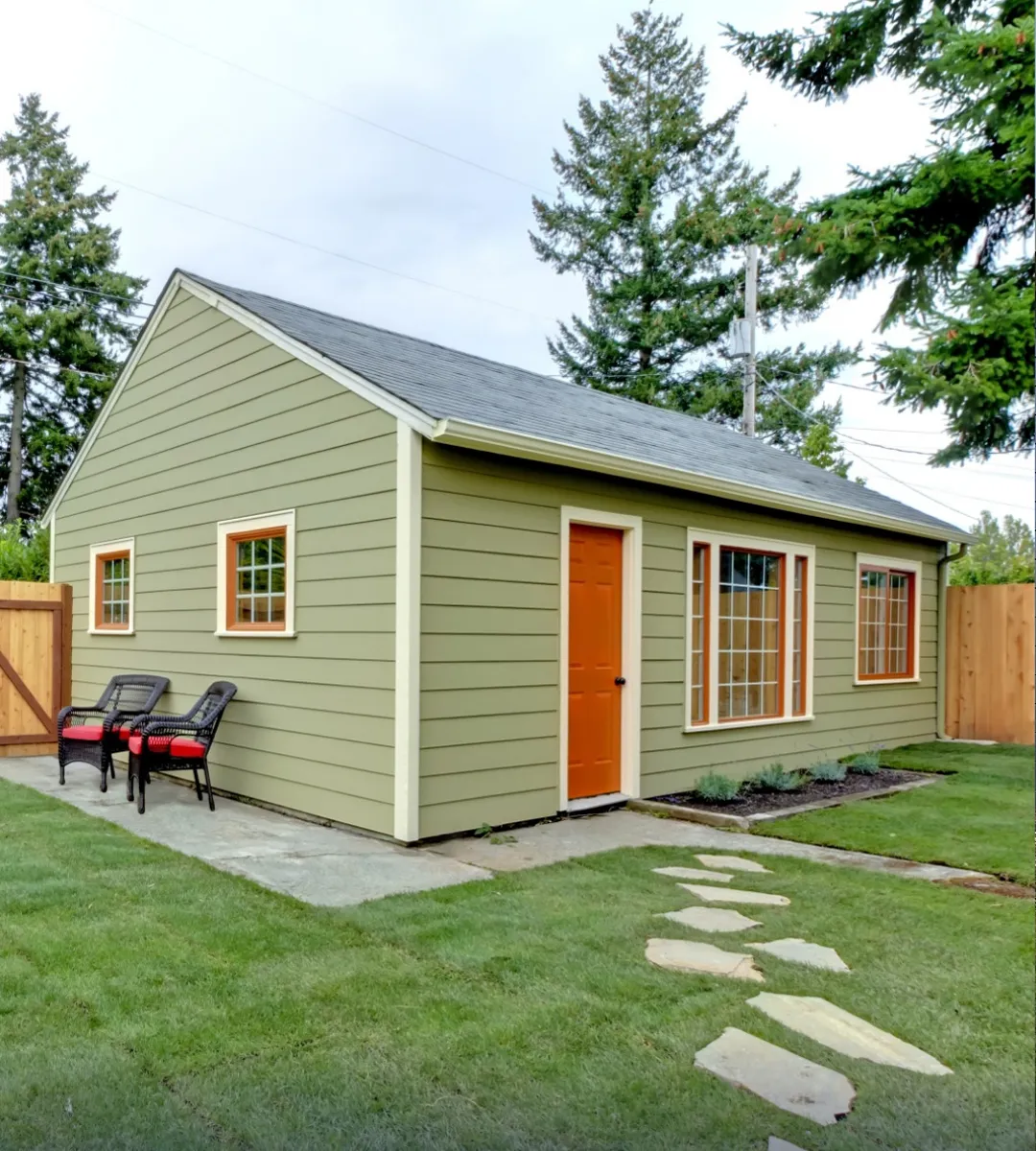 Charming backyard ADU with green siding, orange accents, large windows, and a landscaped yard with a stone pathway.