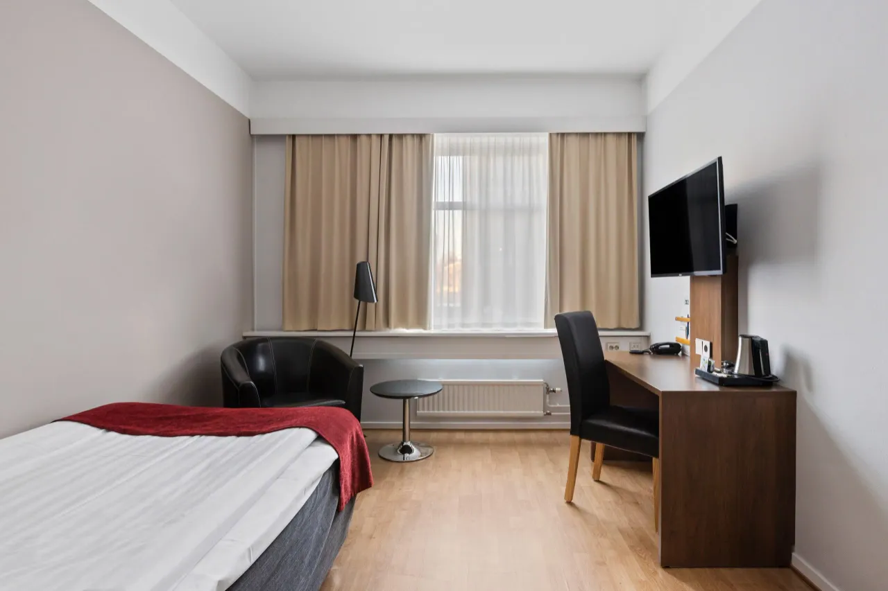 Enkelrum med säng, läderfåtölj, skrivbord och tv framför ett ljust fönster