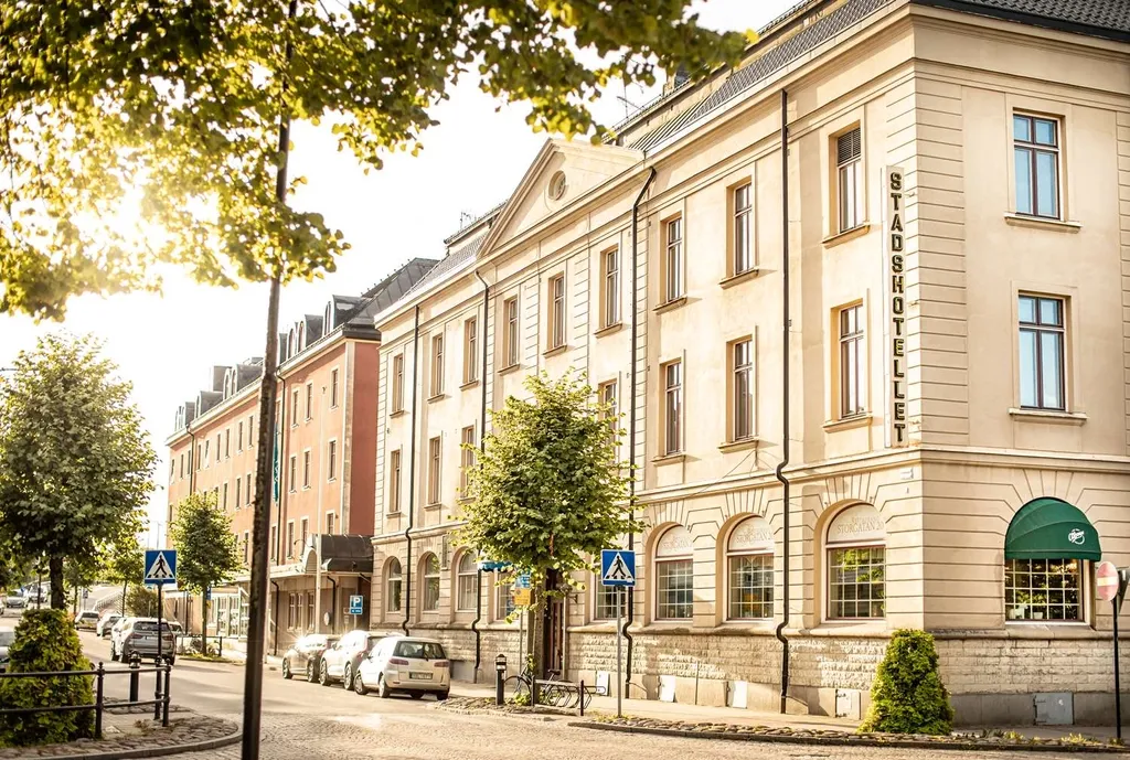 Hotellets ljusa stenfasad med vertikal namnskylt, i solsken bland gröna träd