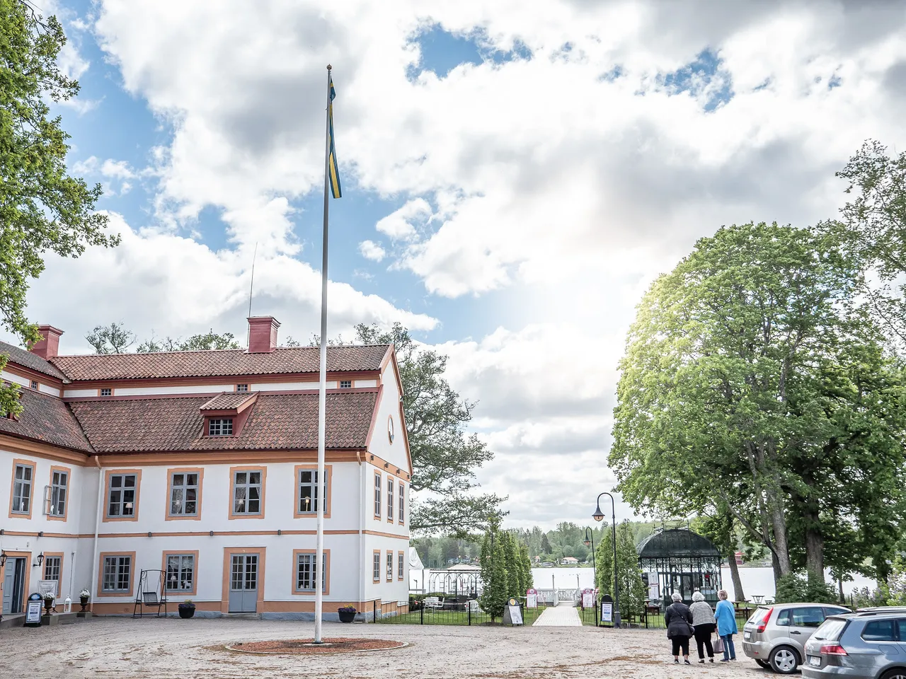 Herrgårdens gårdsplan med flaggstång, den ljusa huvudbyggnaden och lummiga träd