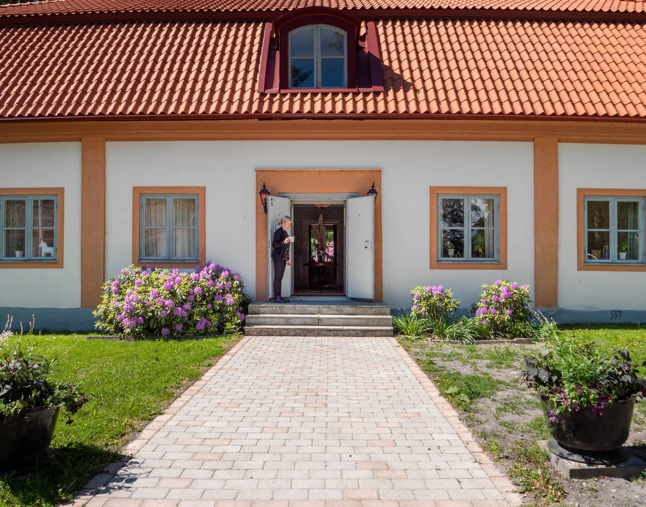 Flygelns ljusa fasad med en stenlagd gång och blommande rhododendron vid entrén