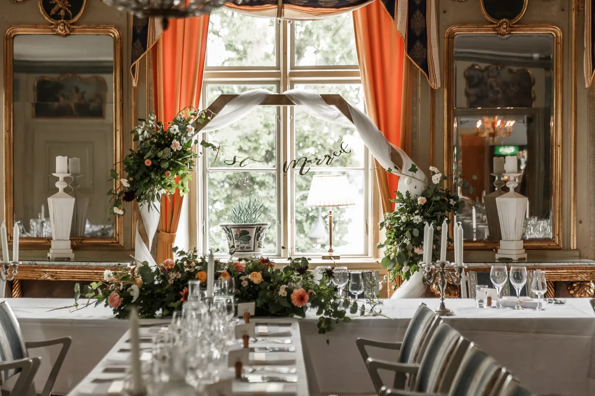 Brudbord med blomsterranka och levande ljus framför ett fönster i en salong med förgyllda speglar
