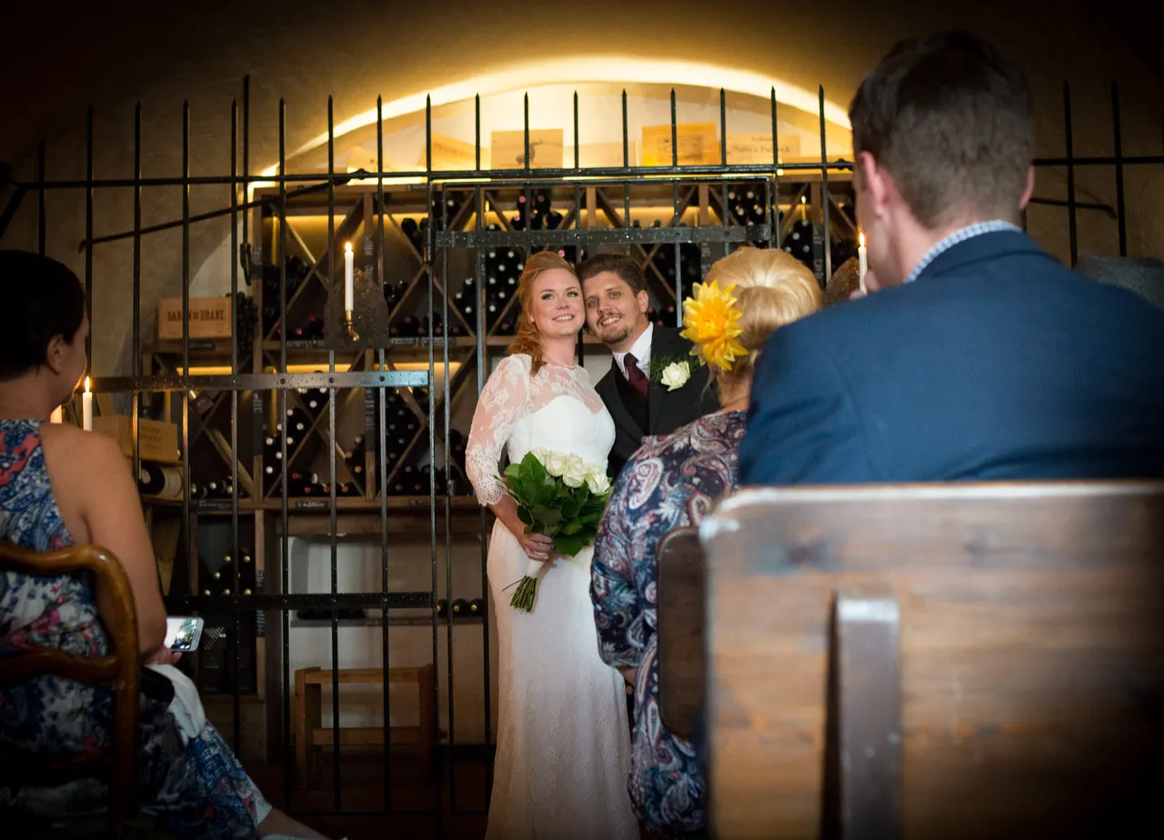 Brudpar framför vinhyllorna i en källare med levande ljus, med gäster på bänkar i förgrunden