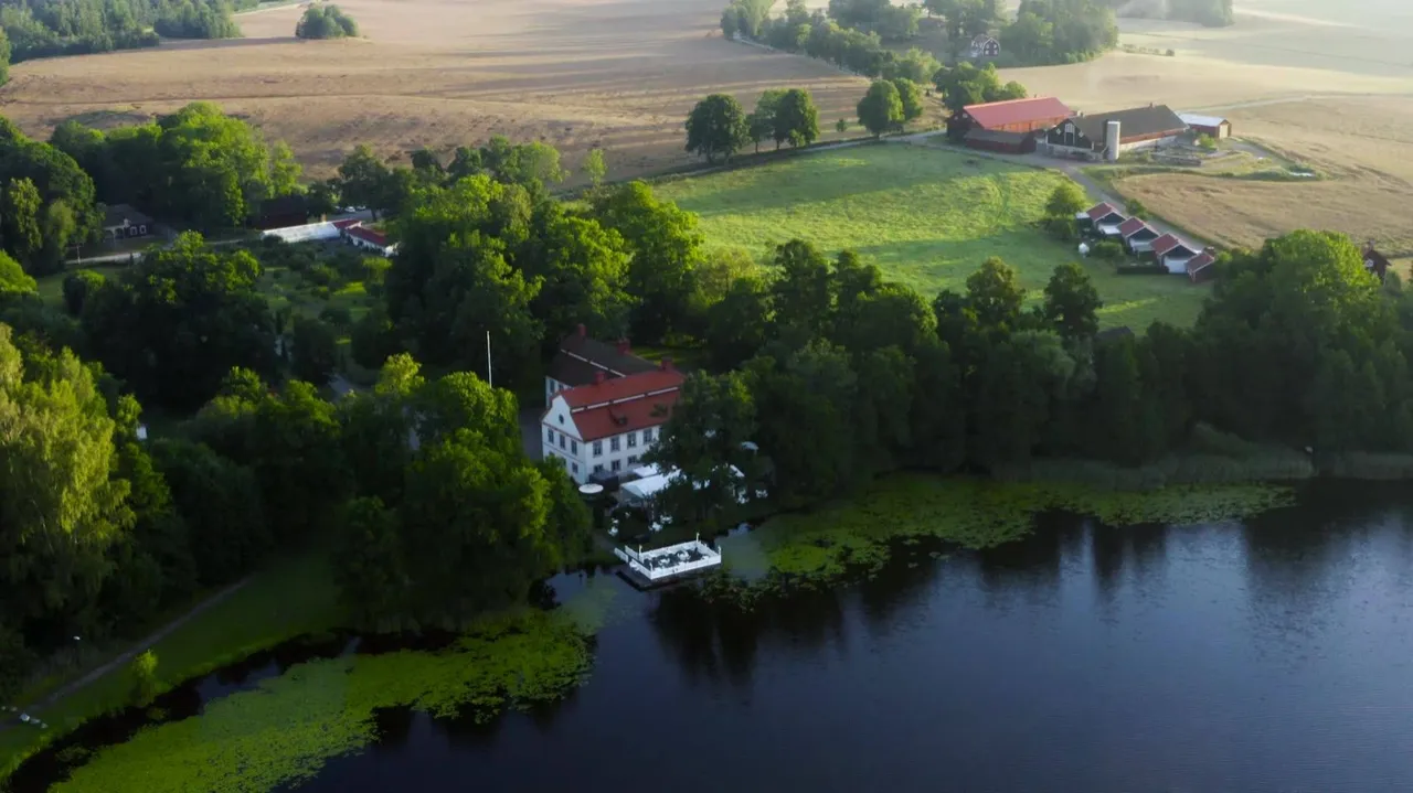 Den vita herrgården med rött tak bland lövträd vid sjön, sedd från ovan en sommarmorgon