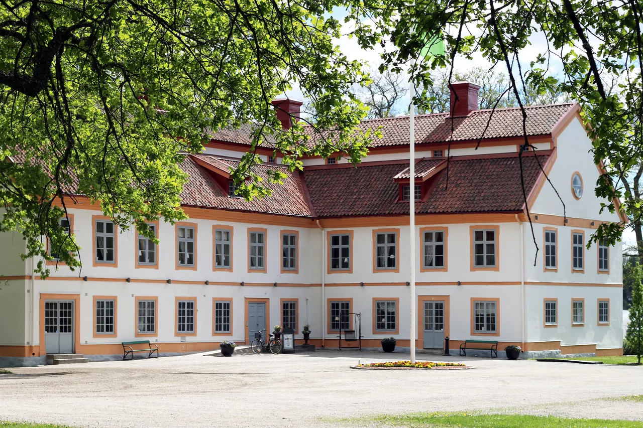 Herrgårdens ljusa fasad med rött tegeltak och blå ytterdörrar, inramad av lövträd en vårdag